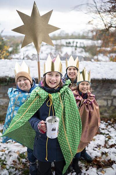 Quelle: © Mika Väisänen / M@RS MediaPortal Sternsinger Vier Kinder mit goldenen Kronen und bunten Umhängen stehen im Schnee, eines hält einen großen goldenen Stern auf einer Stange und eine Spendendose
